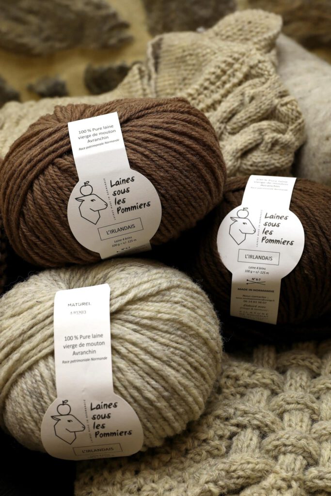 Pelotes de laine de mouton, étiquetées "Laines sous les Pommiers", sur fond de tricot en laine. Durabilité et tradition normande.