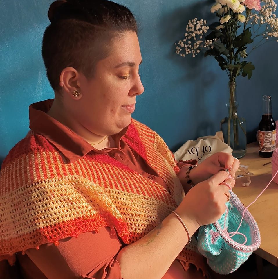 Femme tricotant avec de la laine, vêtue d'un gilet coloré, devant un mur bleu et un vase de fleurs.