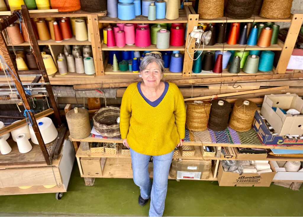 Femme souriante en pull jaune, devant un mur d'enseignes de laine colorées, dans l'atelier de "Laines sous les Pommiers".