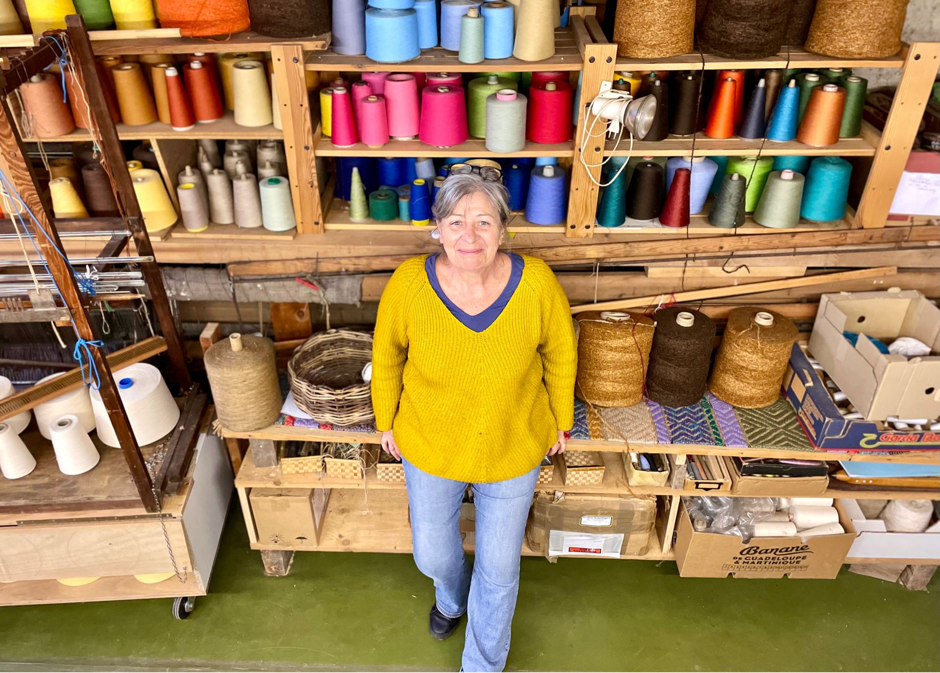 Femme souriante en pull jaune, devant un mur d'enseignes de laine colorées, dans l'atelier de "Laines sous les Pommiers".