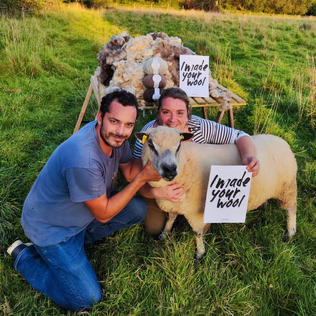 avec un mouton et de la laine, tenant une pancarte "I made your wool" dans un paysage verdoyant.