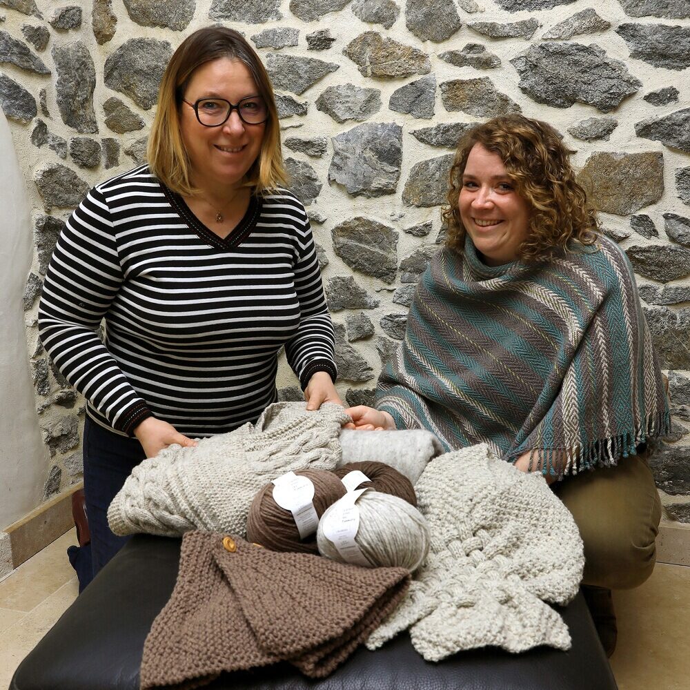 Deux femmes souriantes présentent des vêtements en laine et des pelotes, entourées de produits artisanaux sous un décor de pierres.