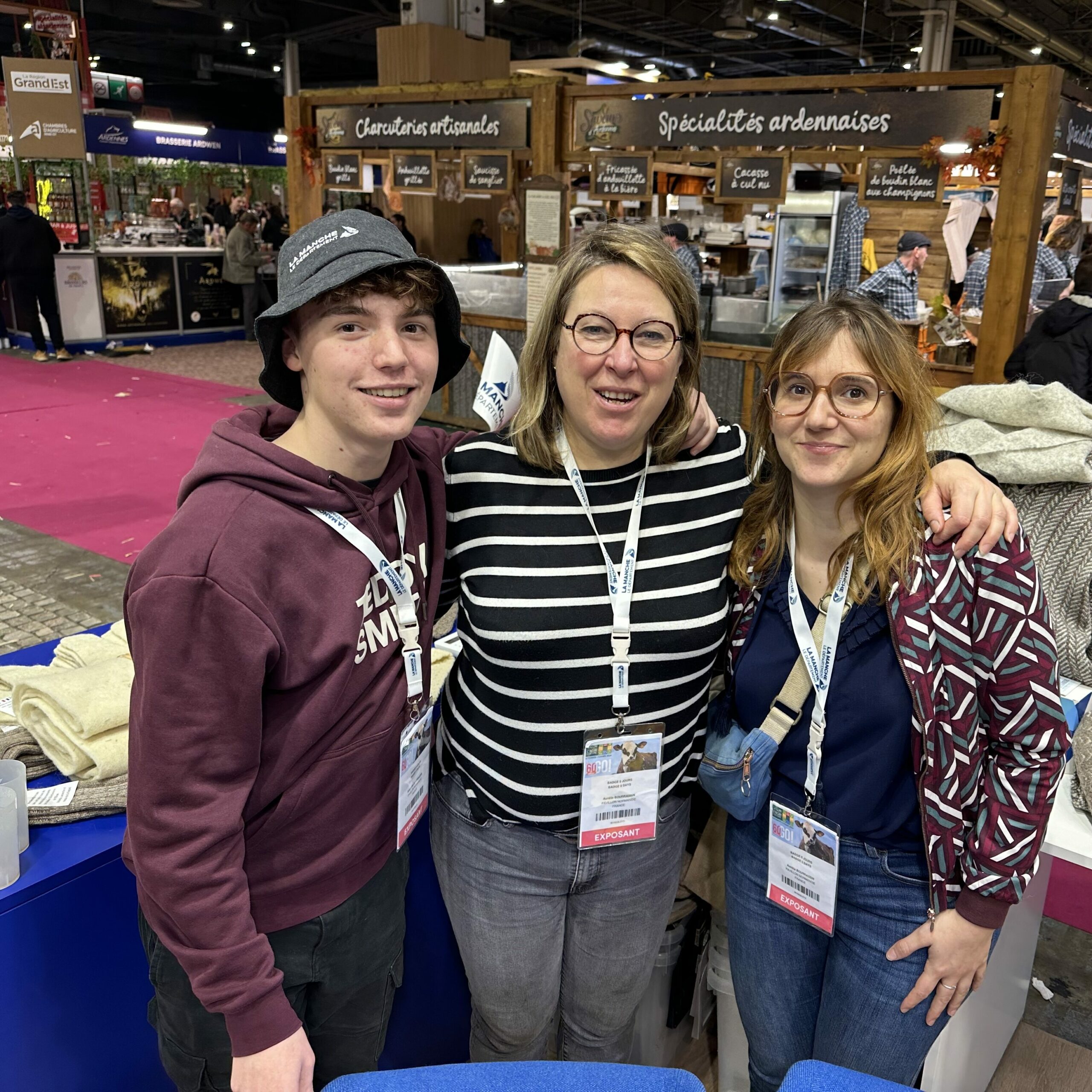 Trois personnes souriantes posent ensemble devant des produits en laine et un stand au salon.