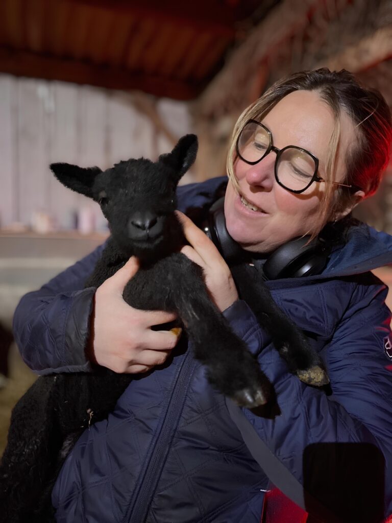 Femme tenant un agneau noir, souriant dans un environnement de ferme, mettant en avant l'authenticité artisanale normande.